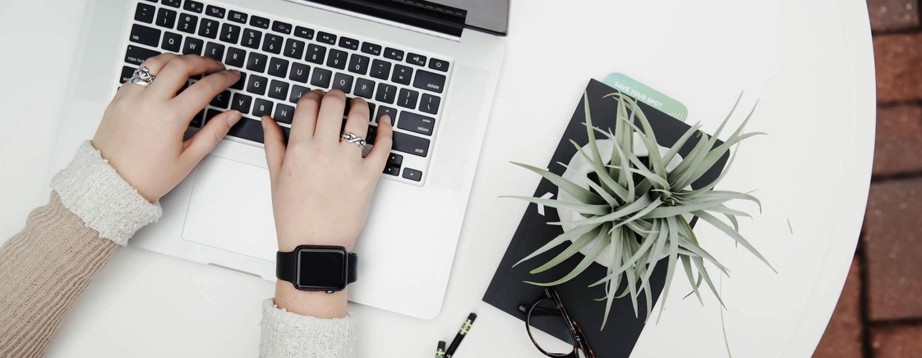 overhead shot of woman working on a laptop at a round table with pencils, her cell phone, glasses, notebook and a potted plant